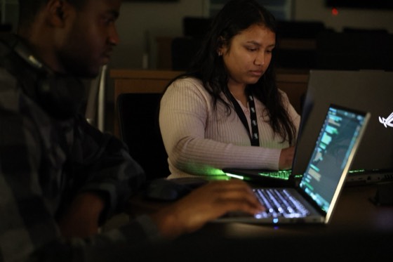 A hacker in a lanyard typing beside a laptop with green keyboard light