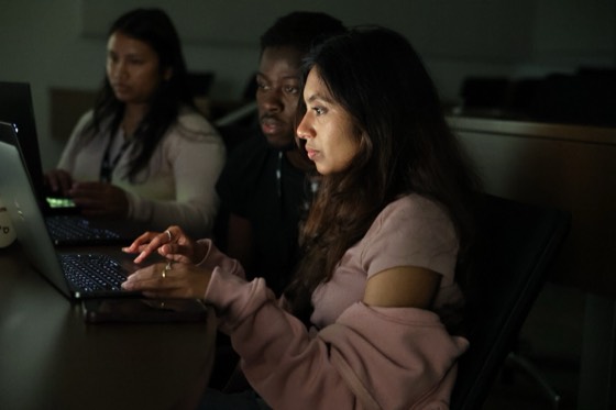 Three hackers lit by their laptop screens in a dark room